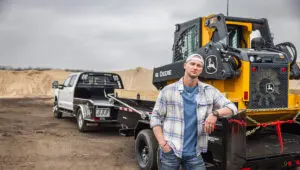 Worker leaning on the Voyager Dump Trailer (DV) with a fully loaded John Deere skid steer