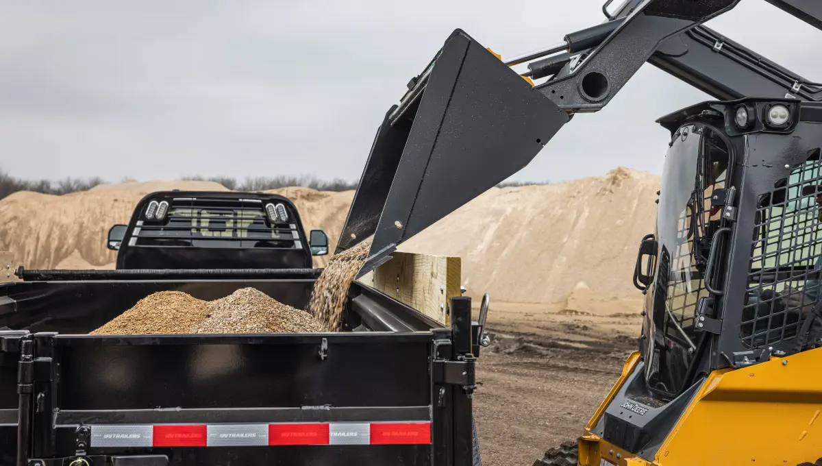 A skid steer loader dumping aggregate material into the Voyager Dump Trailer's bed