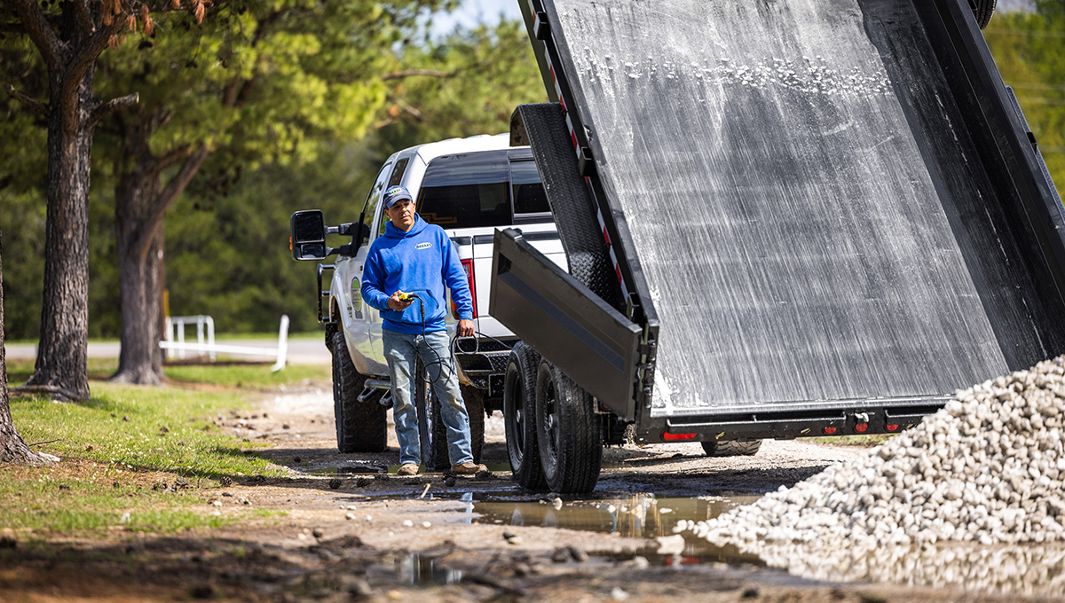 Man dumping gravel from the 14K Low-Profile Dump Trailer onto an unpaved road